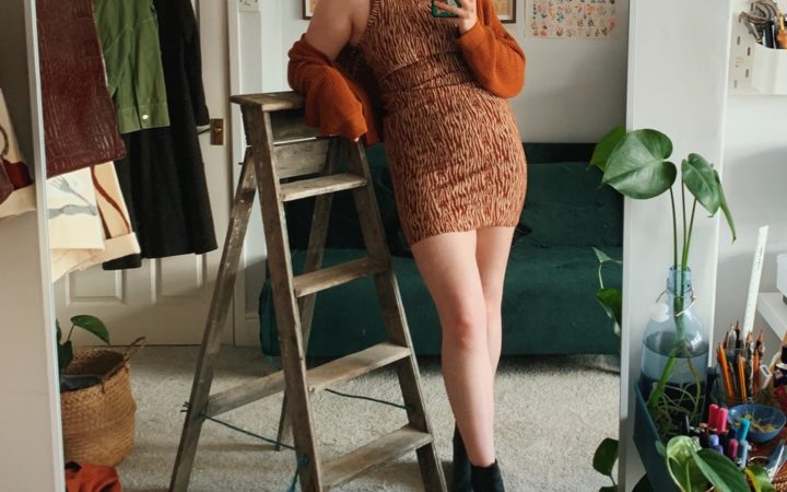 A front view of Roisin in her finished Axis tank dress, styled with a cardigan loosely worn. Roisin is resting an arm on the top of a step ladder which is to the side of her.