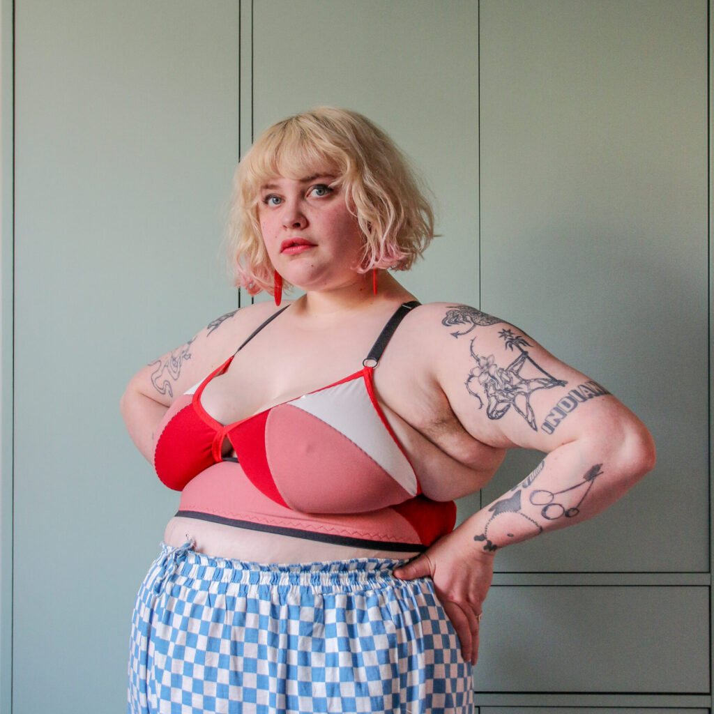 An image of Lydia, a plus sized model with tattoos wearing a colorblocked Ascension Bra in red, pink, white and black. She is standing in front of some wardrobes in a sage green. She is looking directly at the camera, hand on hips in a power pose. 